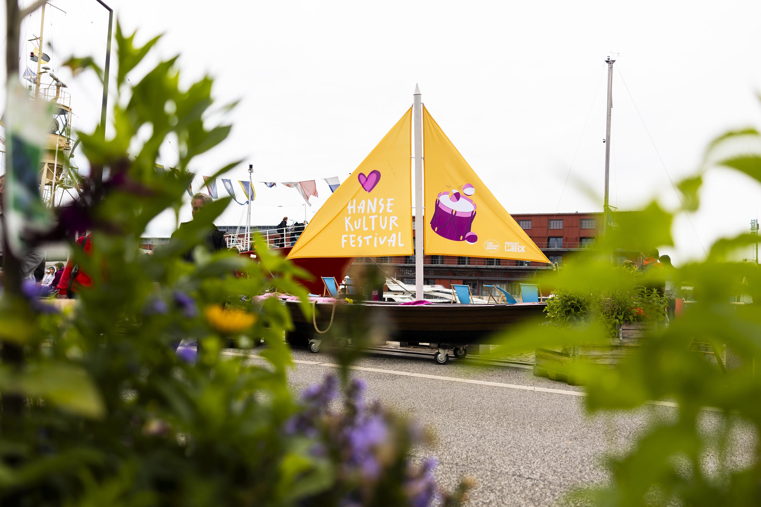 Segelboot mit gelbem HanseKulturFestival-Segel im Lübecker Hafen, eingerahmt von Blumen und Pflanzen.